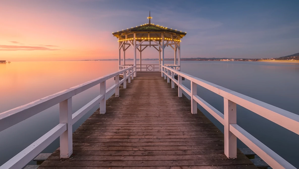 Wooden pier with white railings extending toward a gazebo at sunrise over calm water.