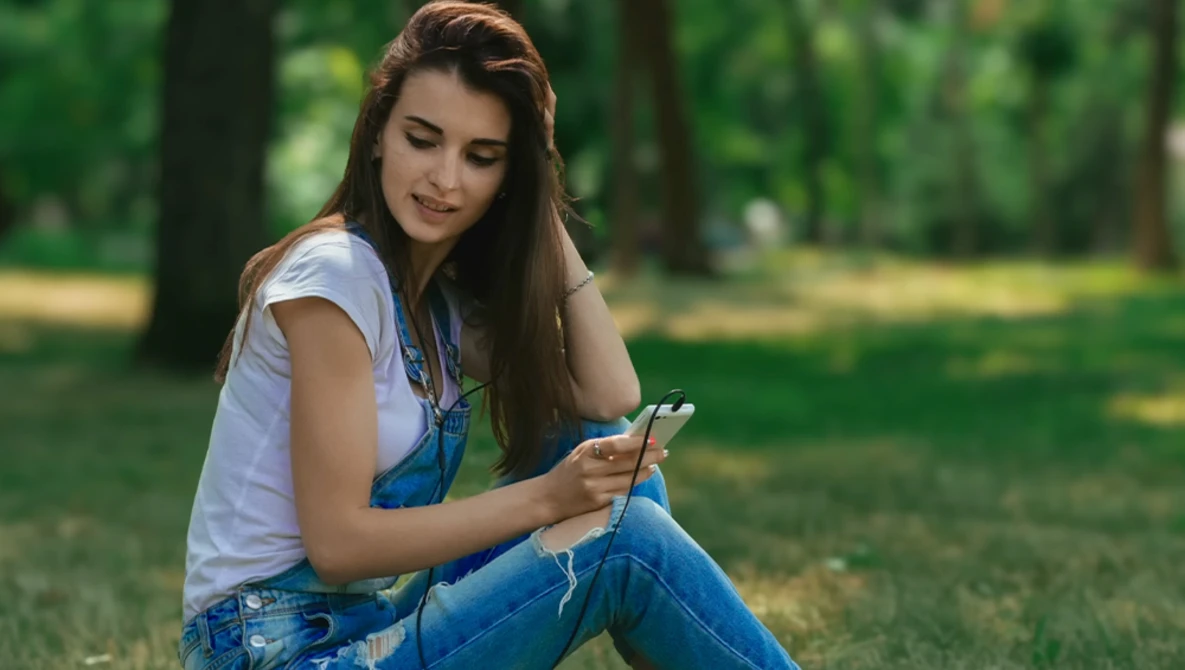 Young woman in denim overalls and white t-shirt sitting outdoors holding a smartphone.