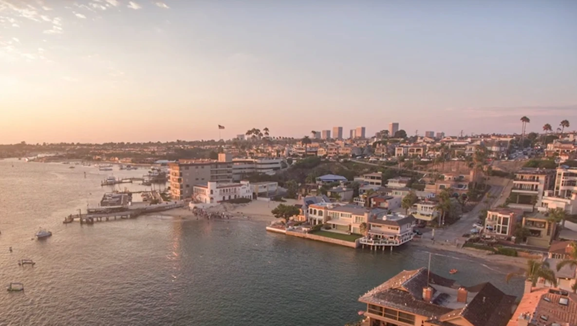 Coastal waterfront neighborhood at golden hour with bay, docks, and distant skyline under clear sky.