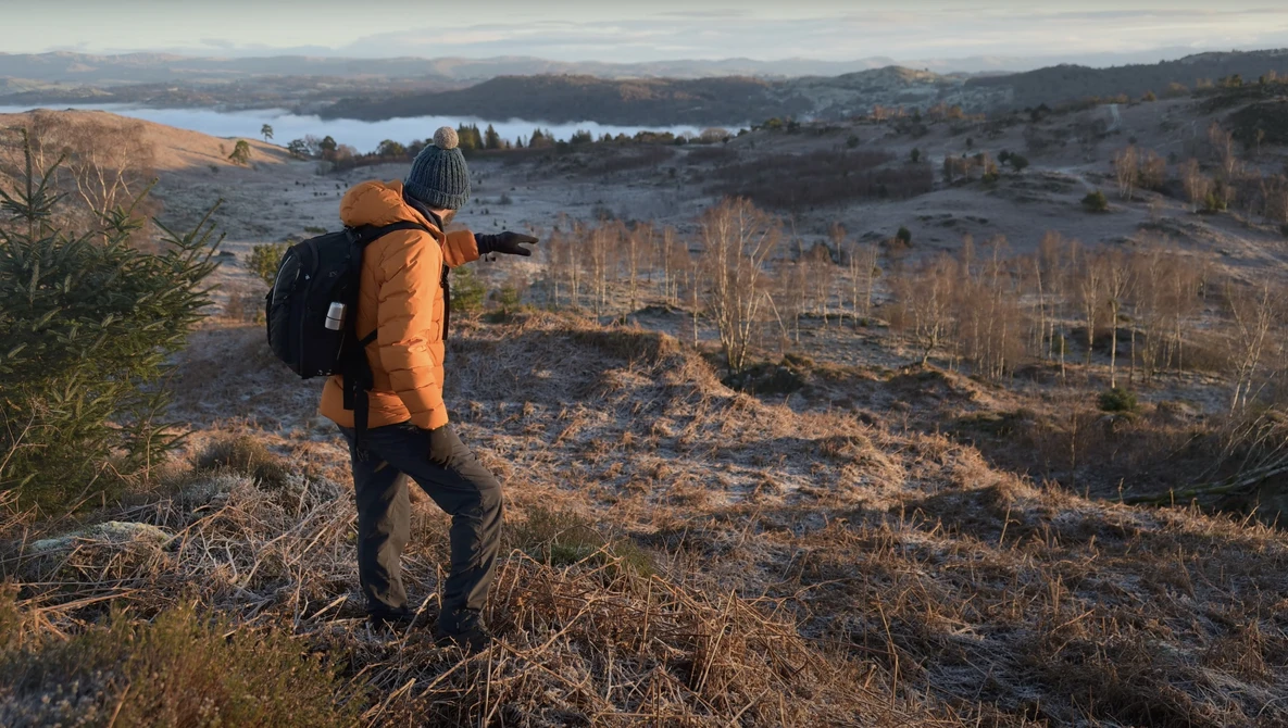 Hiker in orange jacket with backpack overlooking a valley with lakes and bare trees during golden hour.