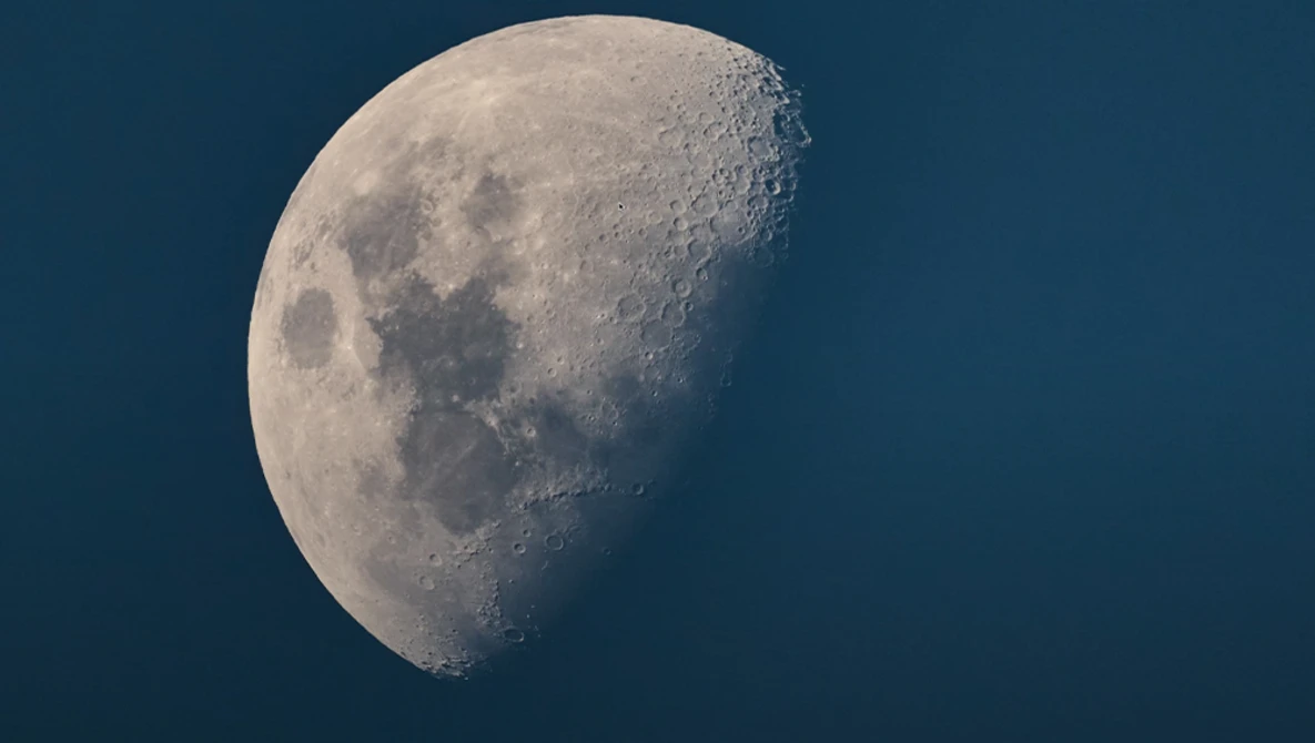 Waxing gibbous moon against dark blue sky showing craters and dark maria in sharp detail.
