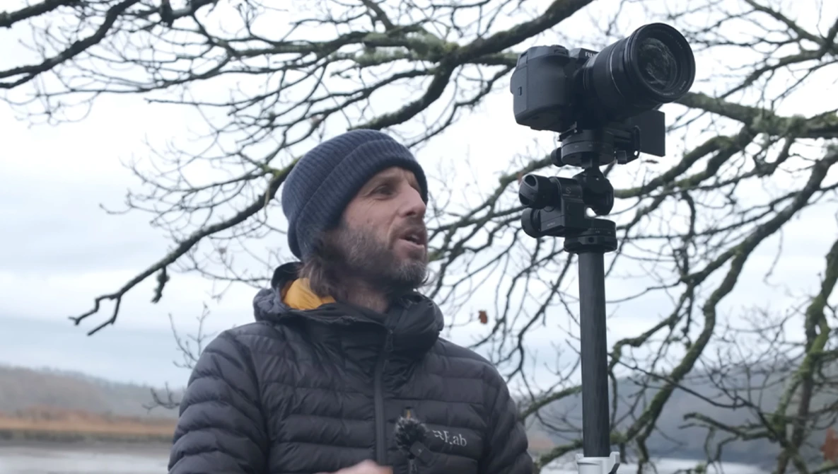 Videographer in winter gear operating a professional mirrorless camera on a tripod overlooking a winter landscape.