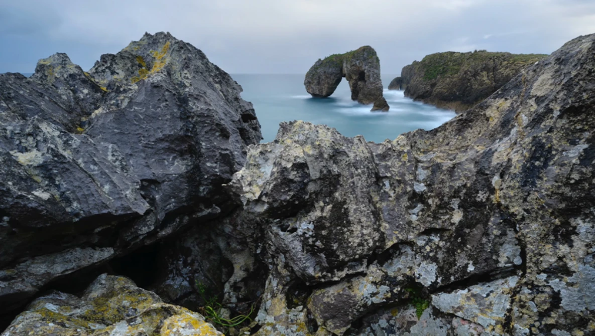 Rugged coastal rock formations with turquoise water and a natural stone arch in the distance.