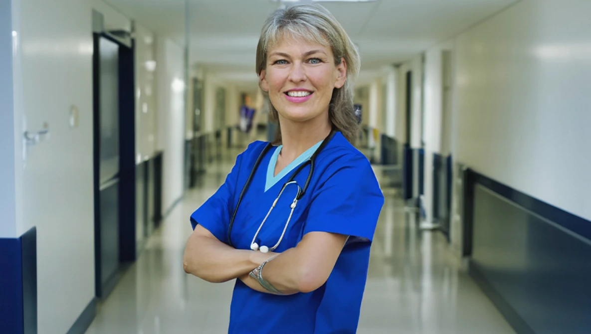 Healthcare professional in blue scrubs with stethoscope, arms crossed in hospital corridor.