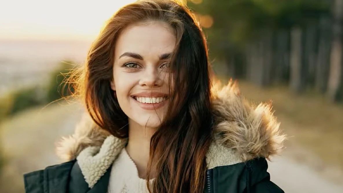 Smiling woman with long dark hair wearing a dark jacket with sherpa-lined collar outdoors during golden hour.