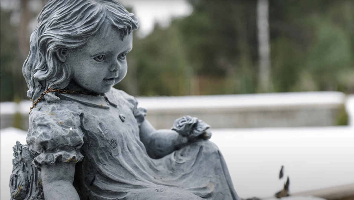 Stone statue of a young child in profile, holding a small flower, with weathered patina.