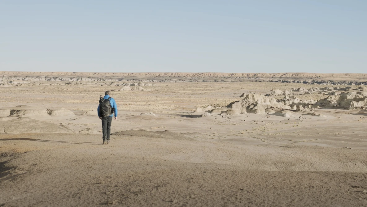 Solitary hiker with blue backpack walking across vast, barren desert terrain scattered with rocks.