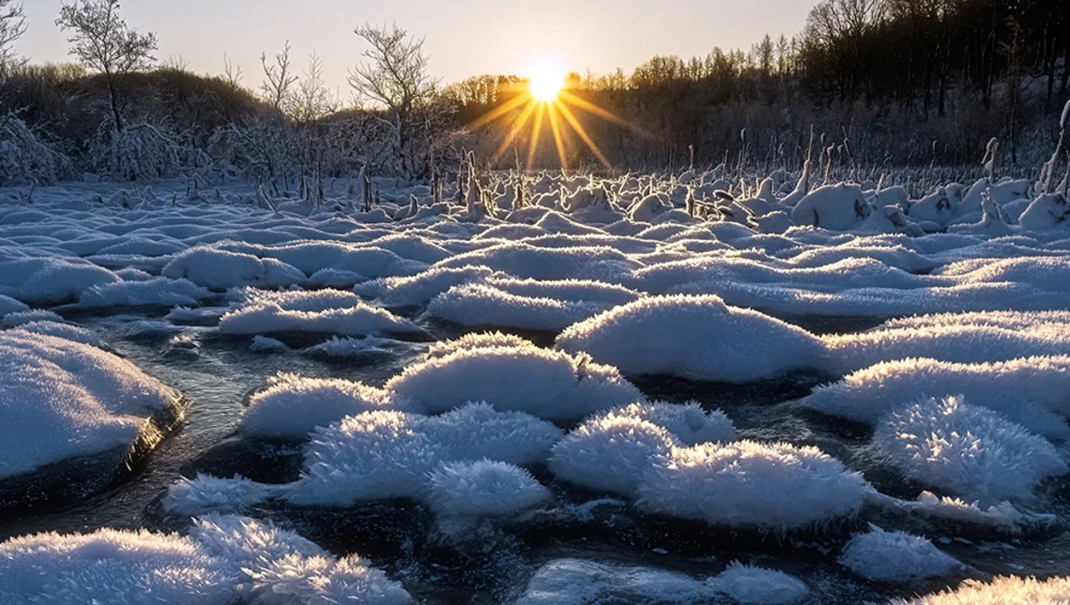 Icy boulders scattered across a partially frozen river at sunrise, backlit by golden sunlight and bare trees.