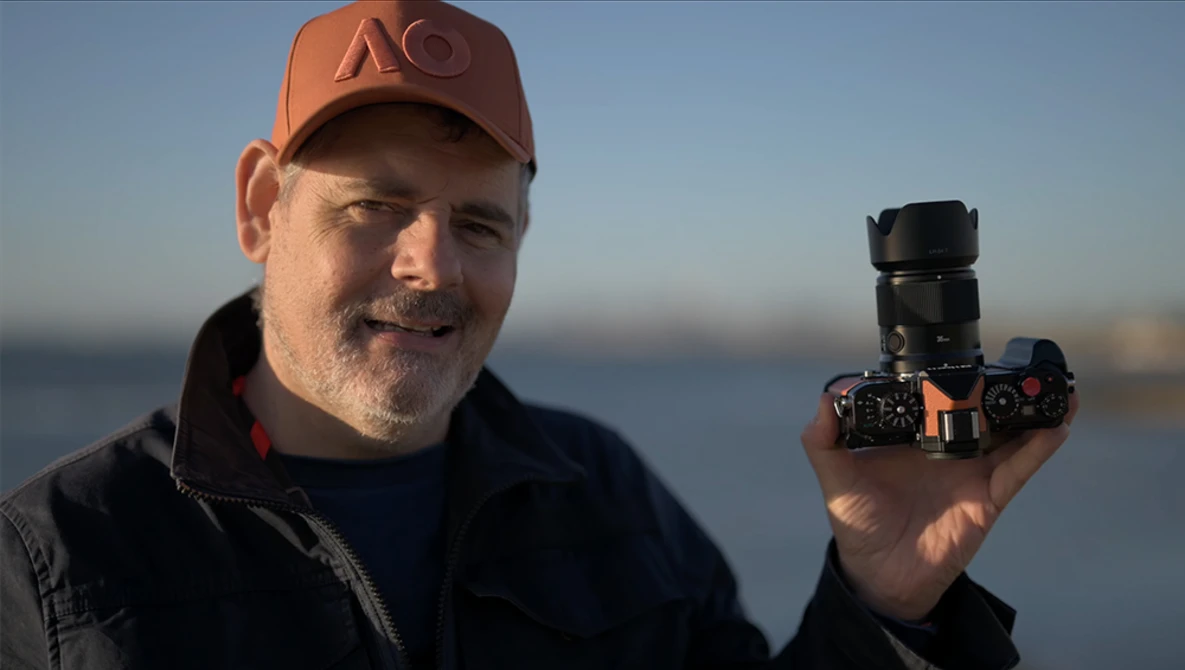Photographer holding a mirrorless camera with telephoto lens against a blurred waterfront backdrop.