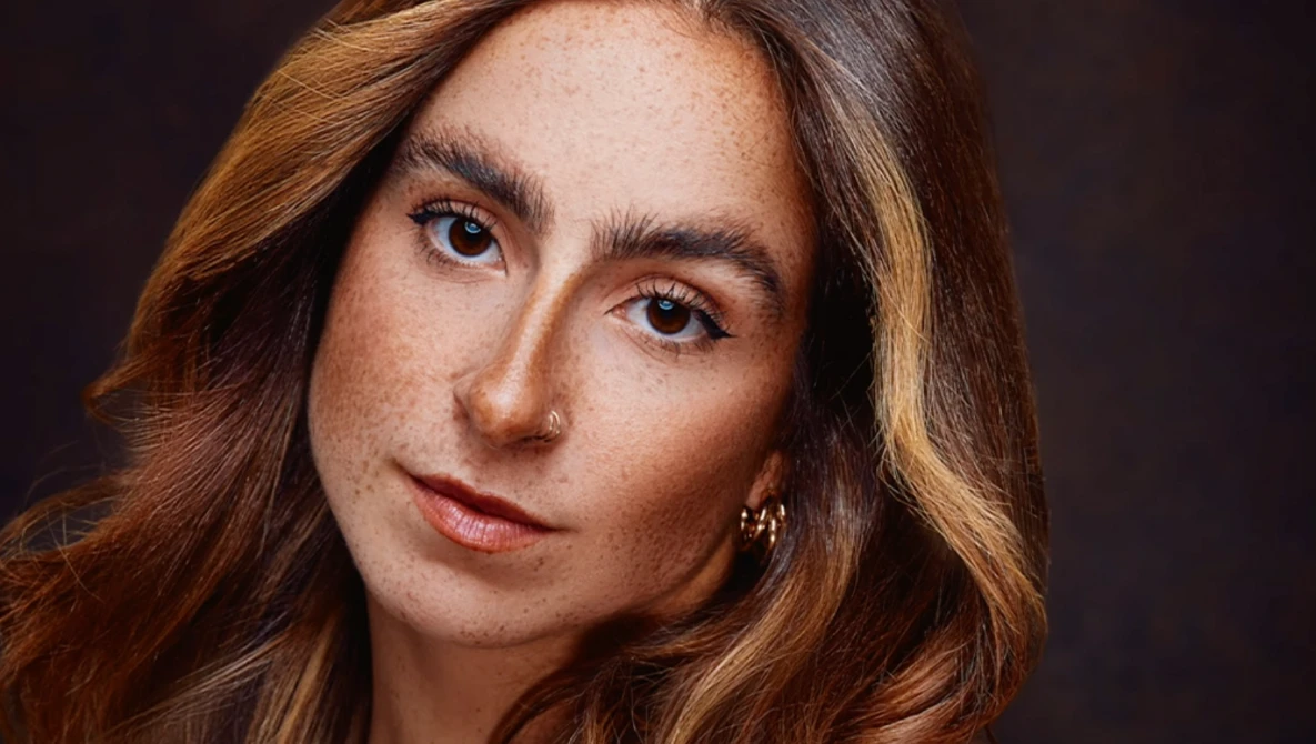 Portrait of a woman with long reddish-brown hair and freckles against a dark background.