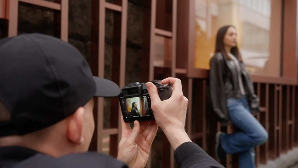Photographer in black cap holding compact camera while framing shot of woman in doorway.