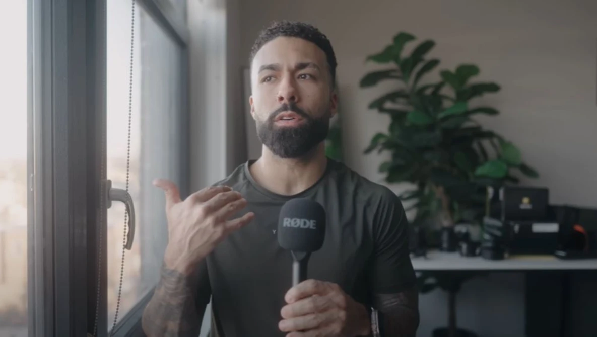Male content creator speaking into a microphone while gesturing toward a window in a studio setting.