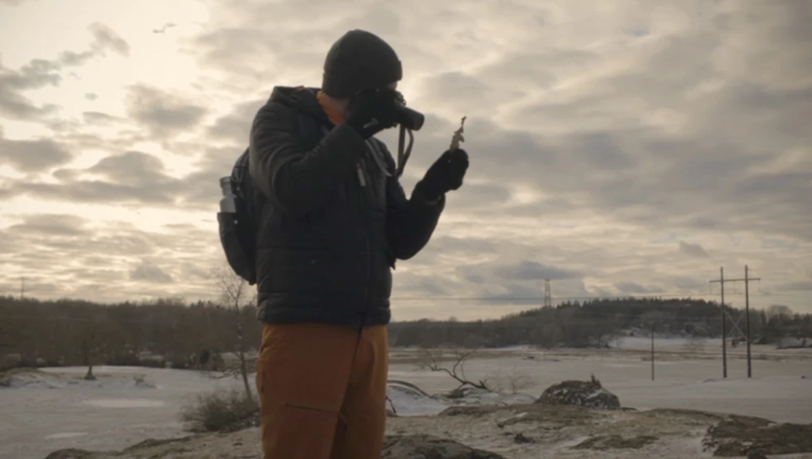 Photographer silhouetted against overcast sky while shooting by a frozen waterway with utility poles visible on distant shore.