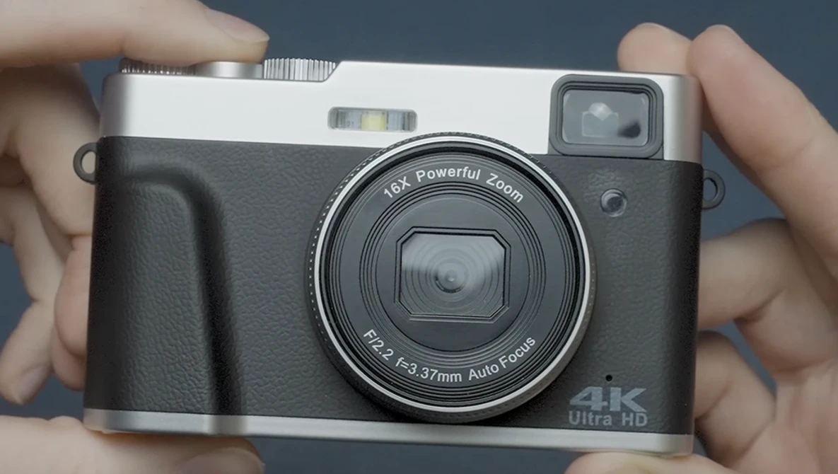 Compact digital camera with black body and silver top plate held in hands against gray background.
