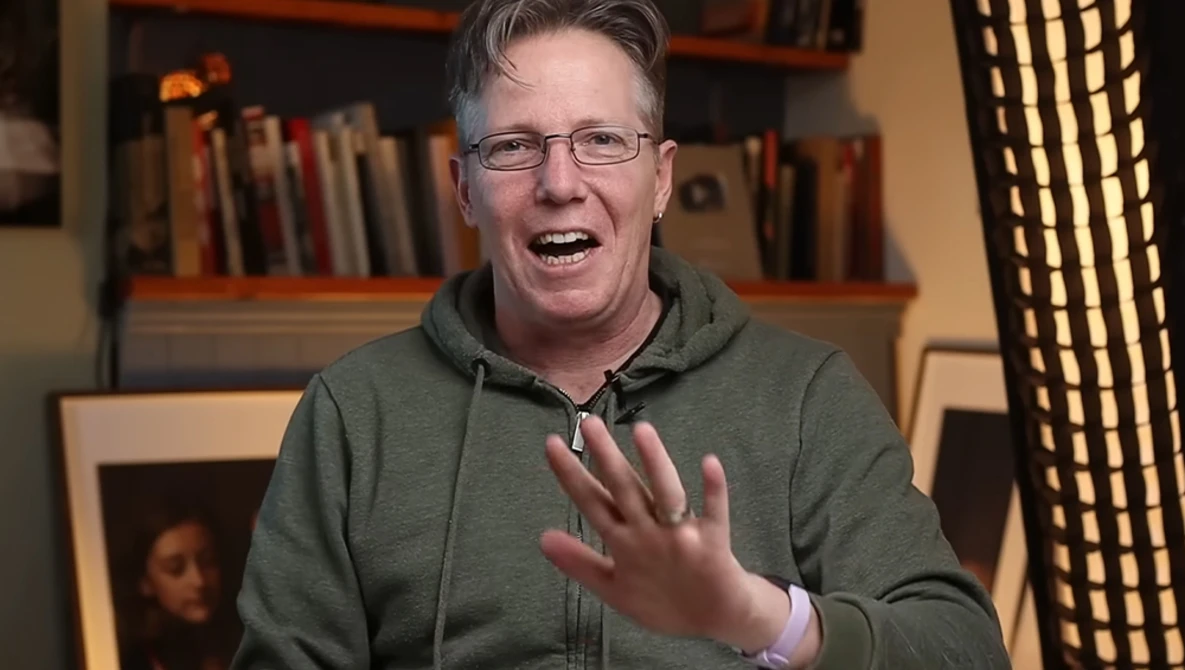 Man in glasses and olive hoodie gesturing while speaking, with bookshelves and warm lighting in background.