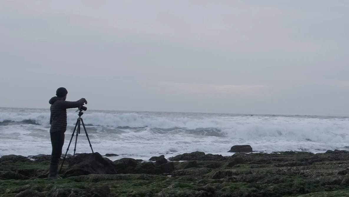 Photographer on tripod positioning equipment on rocky shore facing turbulent ocean waves.