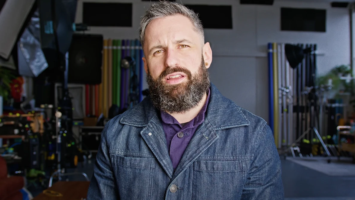 Bearded man with gray hair wearing denim jacket over purple shirt in workshop studio.