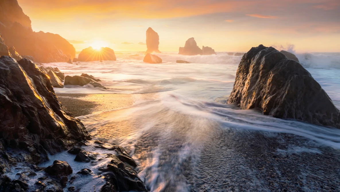Rocky sea stacks silhouetted against golden sunrise over a misty ocean beach.