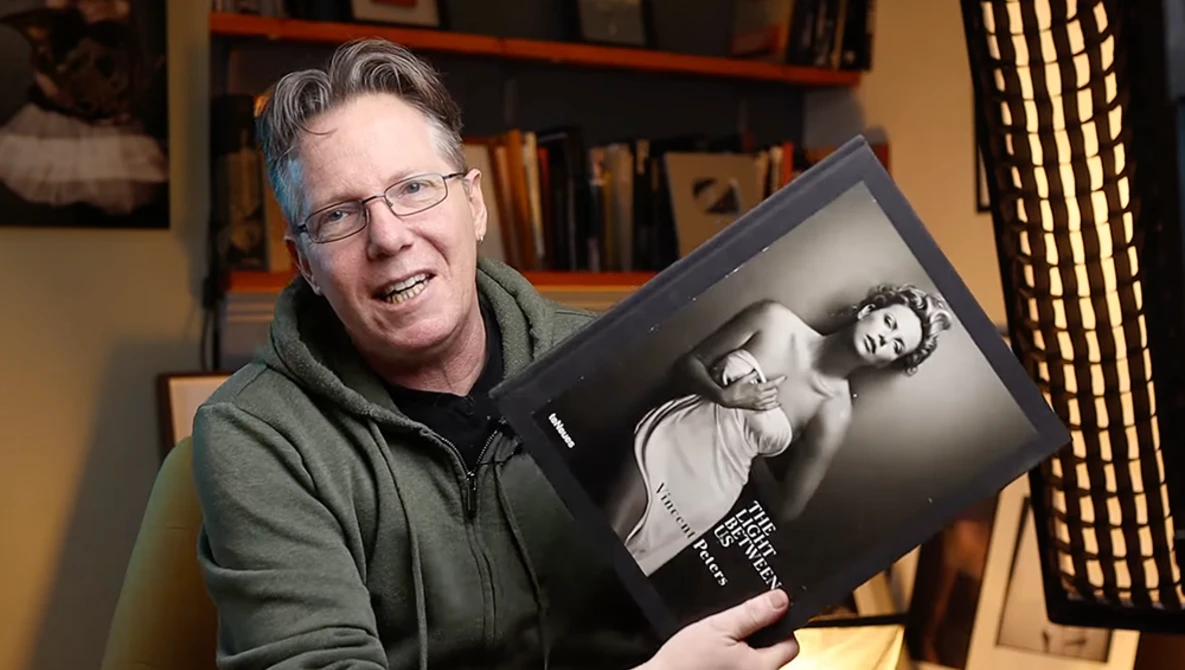 Photographer holding a black and white portrait book in a studio setting with bookshelves behind.
