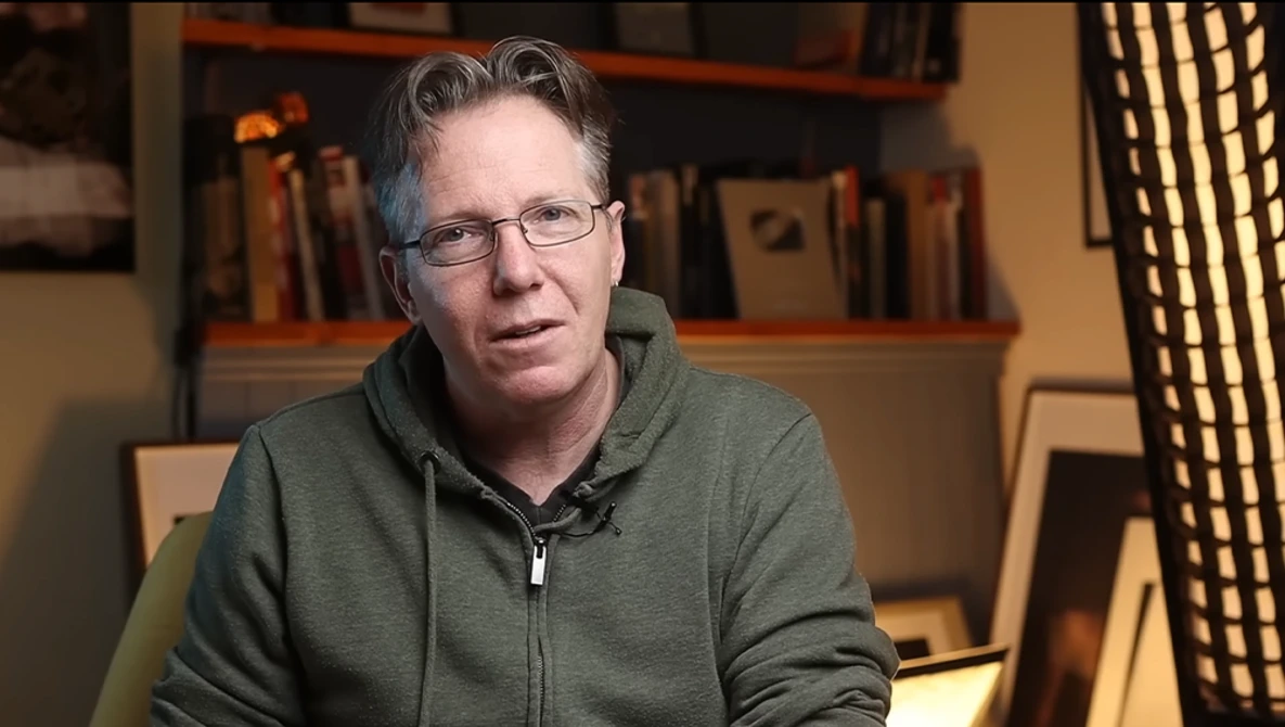 Man wearing glasses and gray zip-up hoodie speaking in an indoor setting with bookshelves behind him.