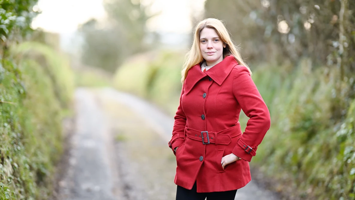 Woman in red coat standing on tree-lined path, hands in pockets, looking at camera.