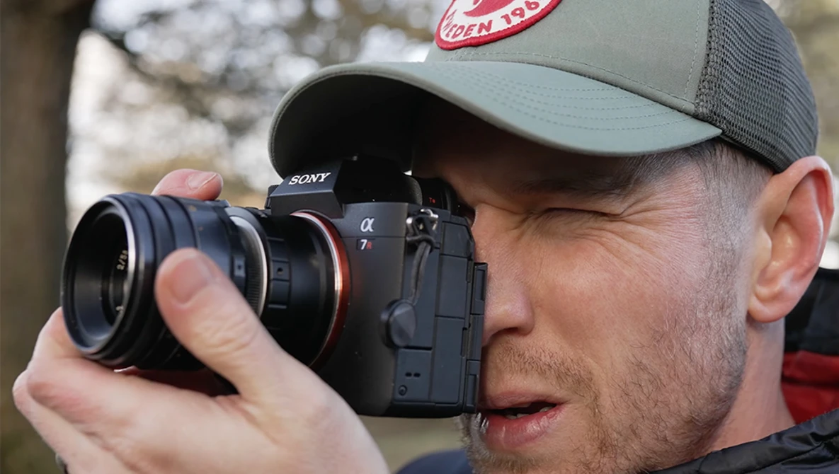 Photographer holding a Sony mirrorless camera with a telephoto lens, wearing a gray baseball cap.