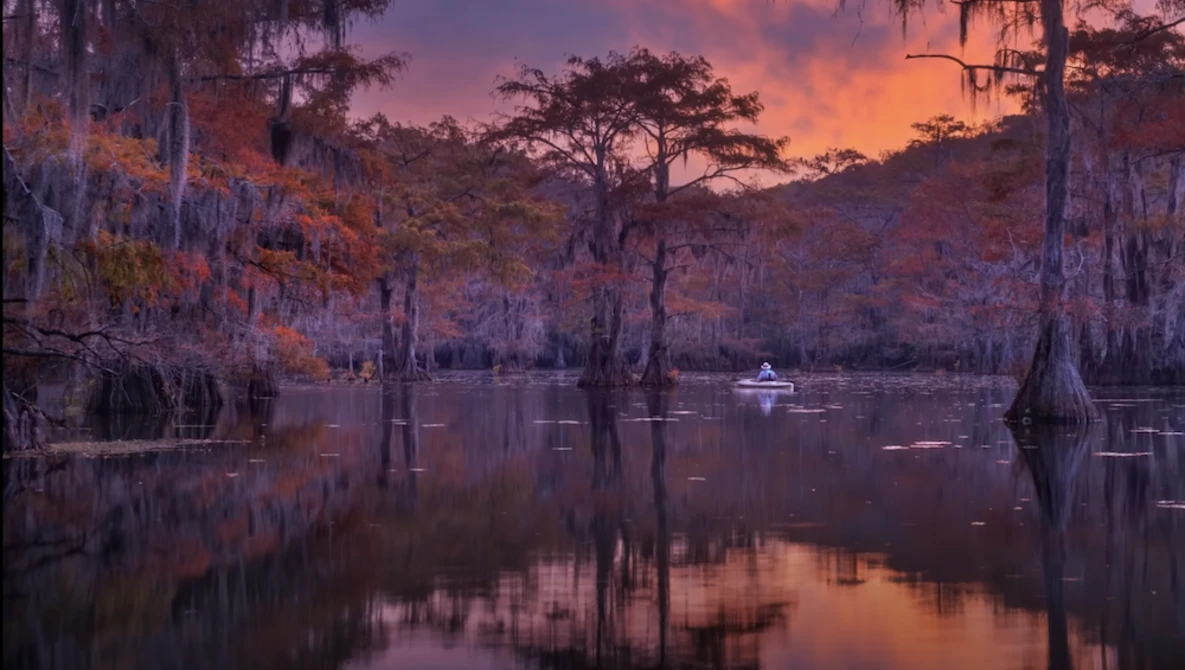 Cypress swamp at sunset with Spanish moss draped trees reflected in still water, small boat in distance.