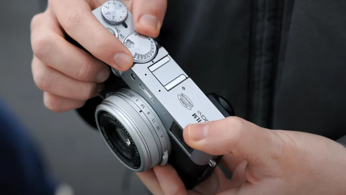 Hands holding a compact rangefinder camera with manual dials and a fixed lens.