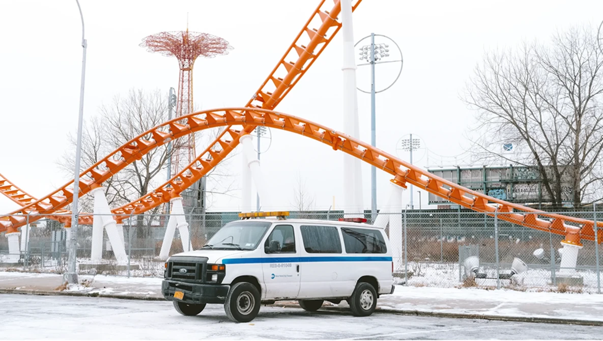 White and blue van parked in front of an orange roller coaster structure in an amusement park setting.