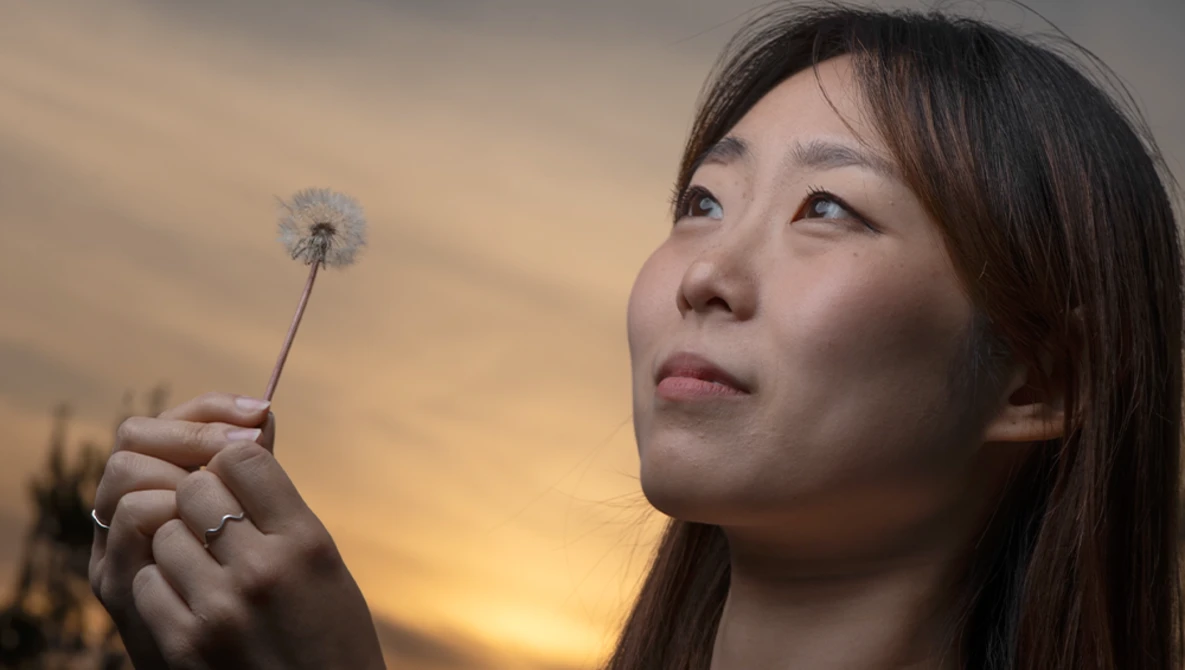 Woman holding a dandelion seedhead against a golden sunset sky.