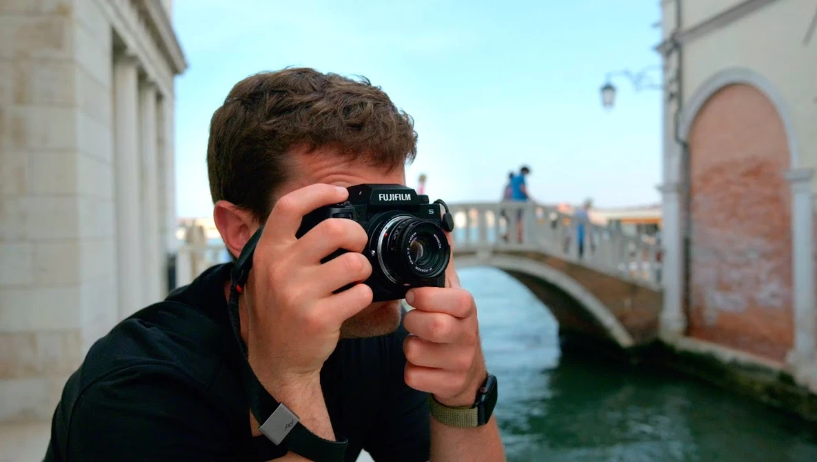Photographer holding a Fujifilm medium format camera up to their face while standing in a European waterside alley.
