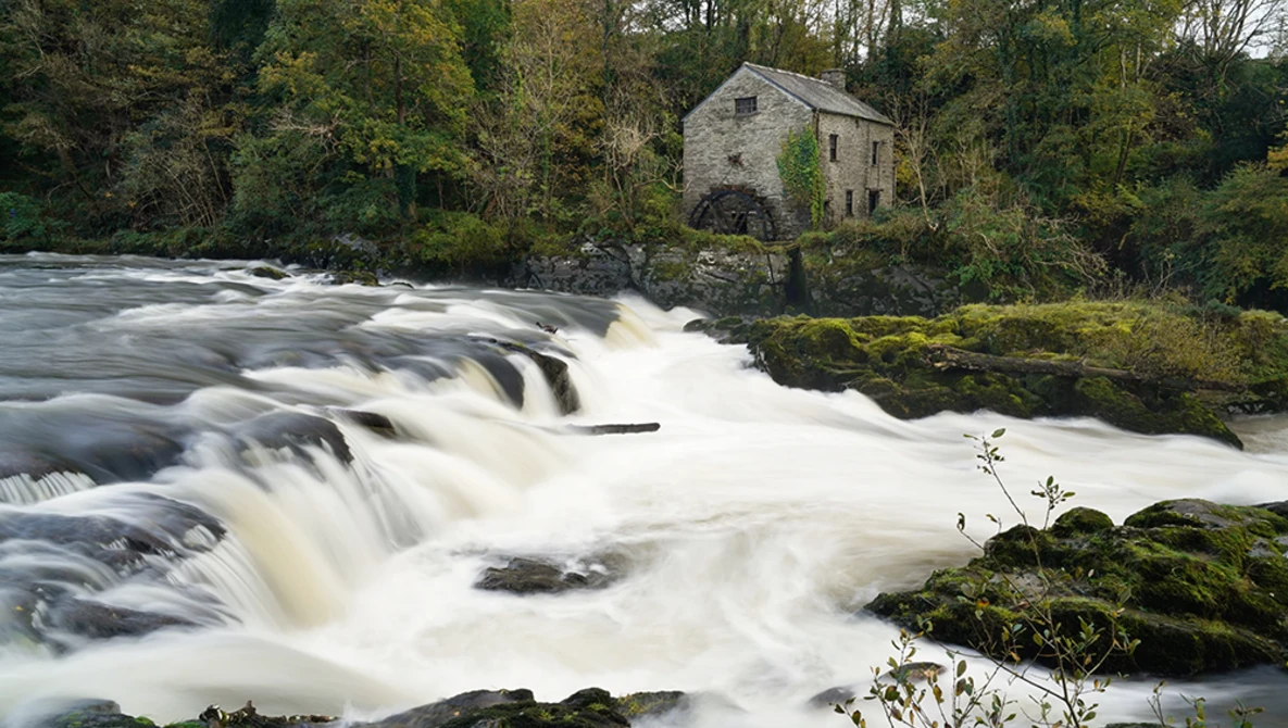 Cascading waterfall with stone mill building surrounded by trees on riverbank.