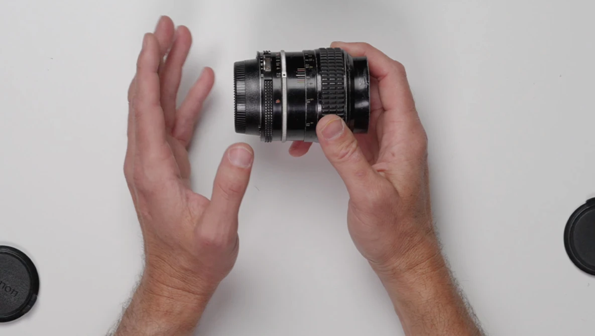 Hands holding a vintage camera lens with focus and aperture rings against a white background.