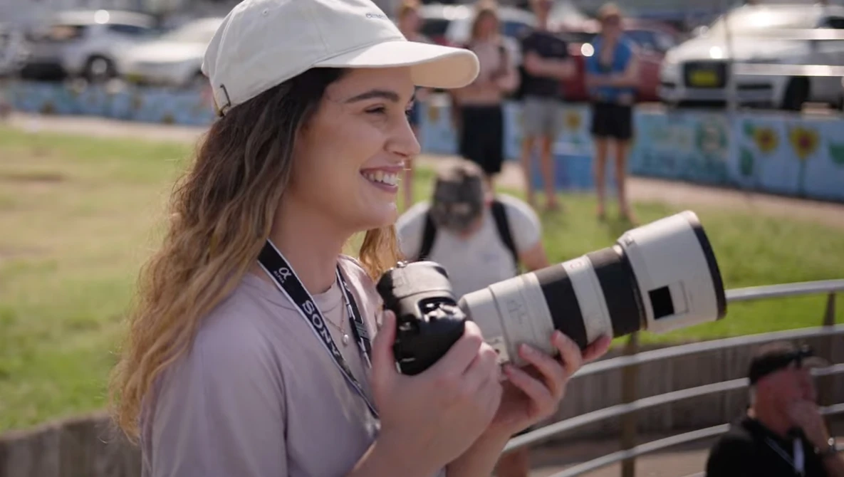Female photographer holding a telephoto lens at an outdoor sports event.