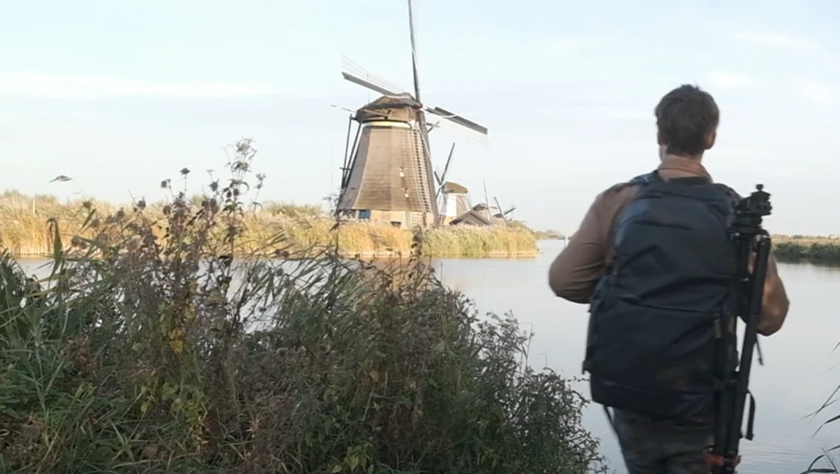 Photographer with camera and backpack standing on grassy bank overlooking traditional Dutch windmill across still water.