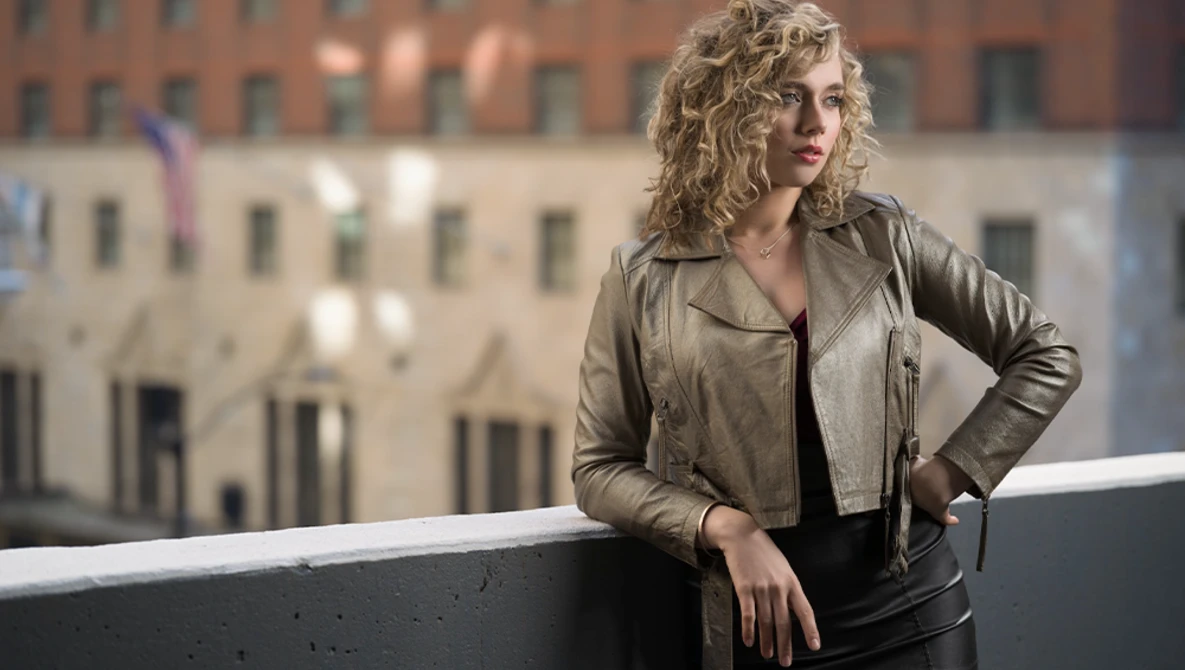 Woman with curly blonde hair leaning against a concrete railing overlooking urban buildings.