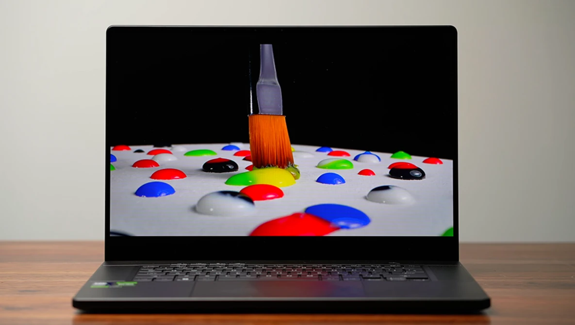 Screenshot of a paintbrush with orange bristles positioned above colorful paint droplets on a white surface displayed on a laptop screen.