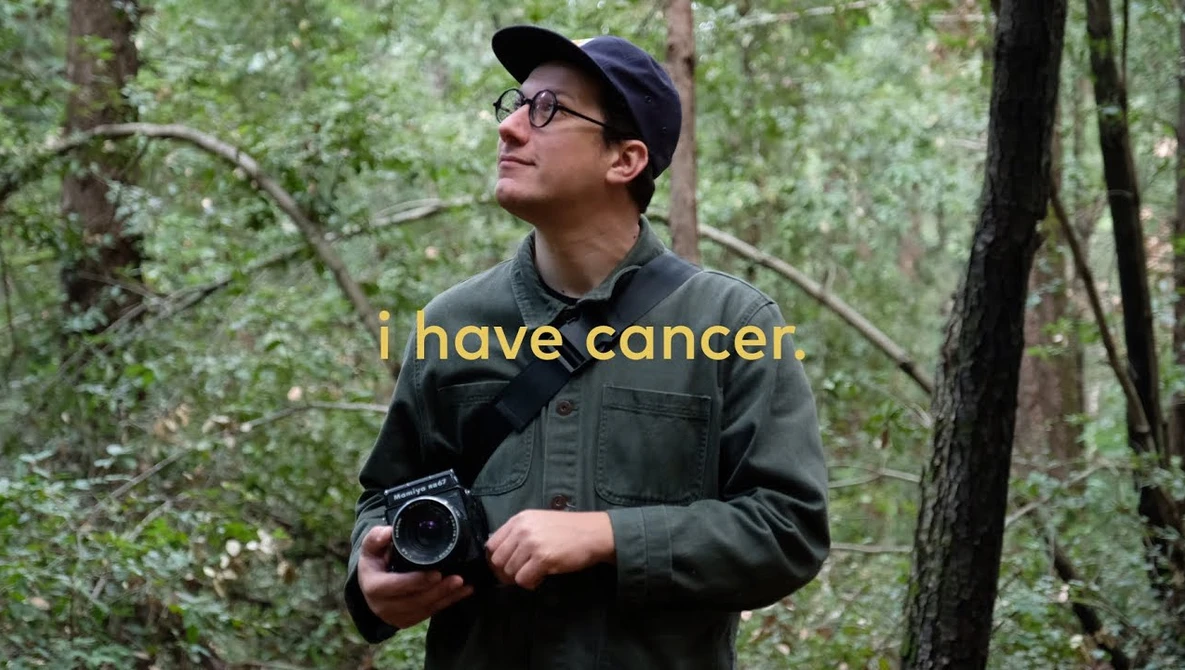 Person in forest holding vintage camera, wearing glasses and dark jacket.