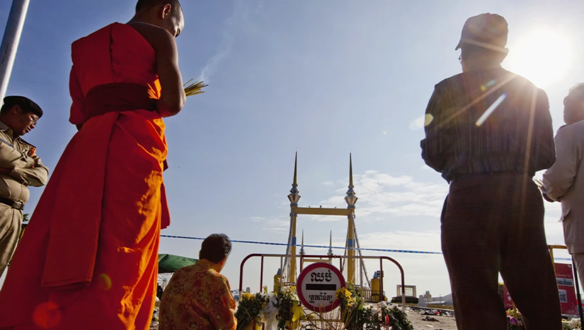 Buddhist monks in red and gold robes stand near a bridge with golden spires during a daytime ceremony.