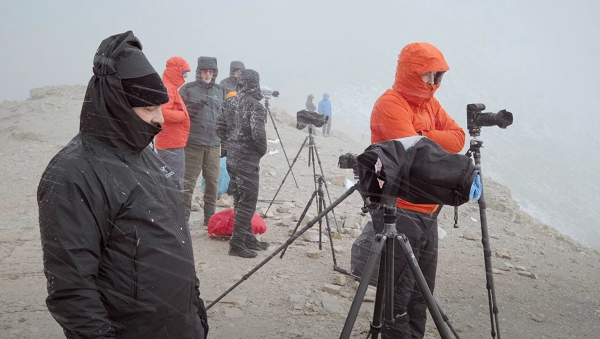 Photographers with telephoto lenses on tripods in heavy fog on a misty mountain ridge.