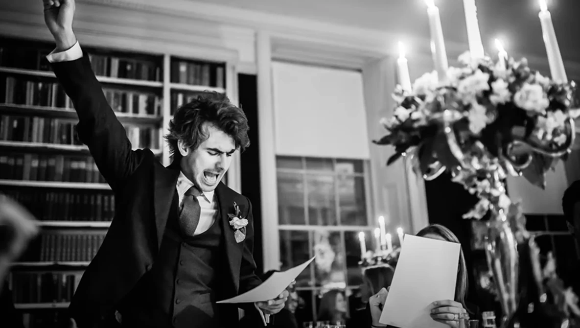 Groom in black tuxedo raising his fist triumphantly while holding papers in an elegant library setting.