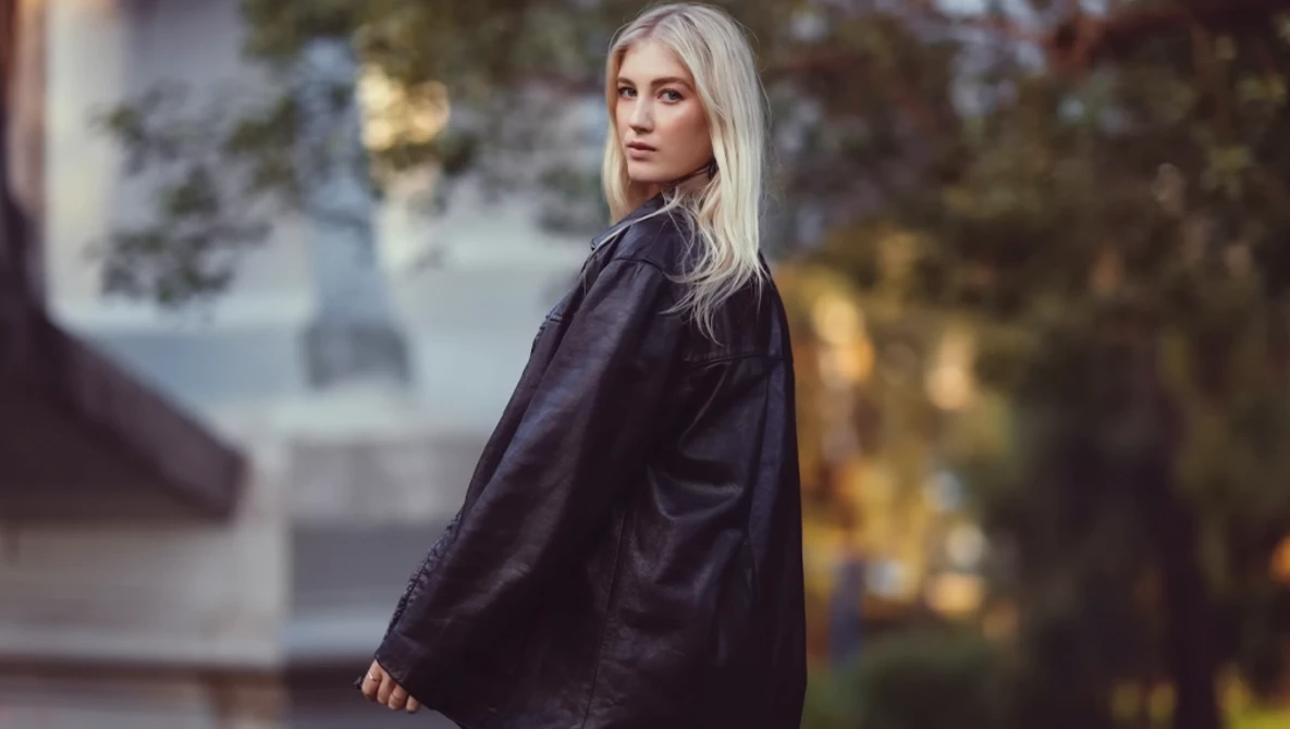 Woman with long platinum blonde hair wearing a dark navy jacket, photographed outdoors in soft golden-hour light.