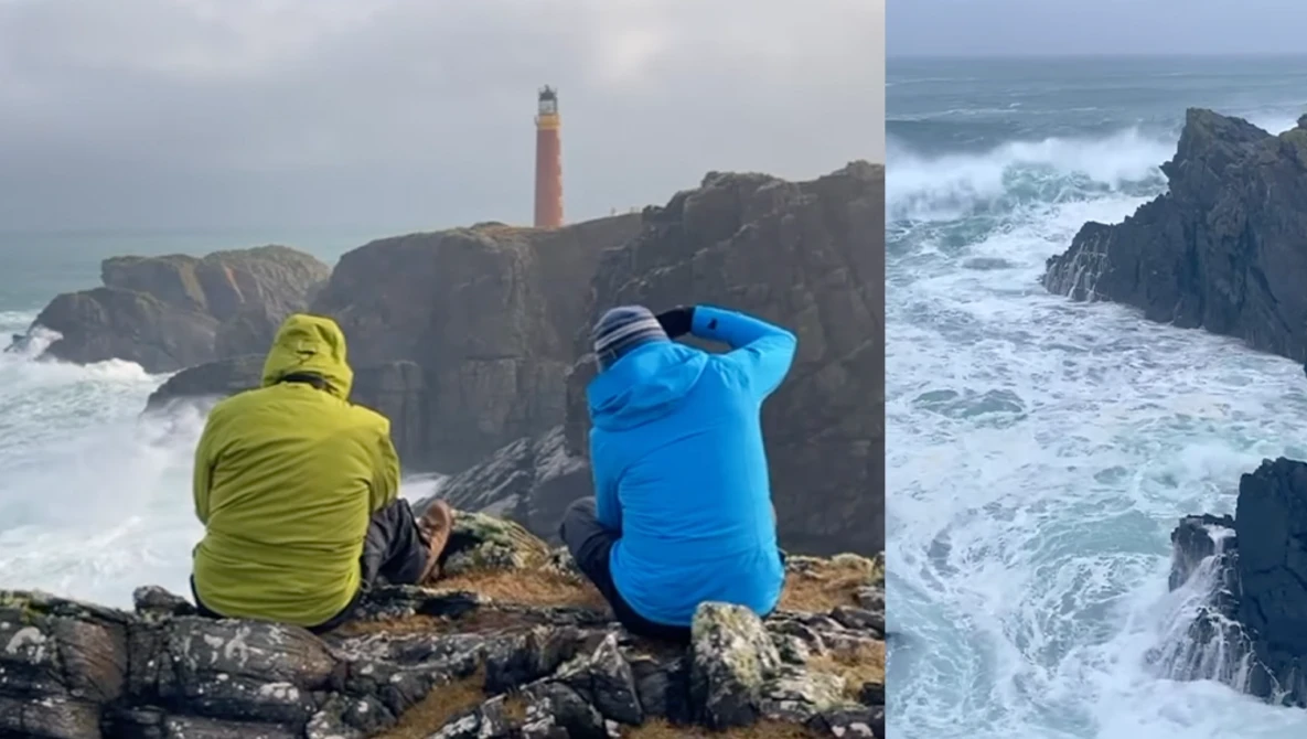 Two photographers in bright jackets observe a dramatic rocky coastline with a red and white lighthouse and turbulent ocean waves.
