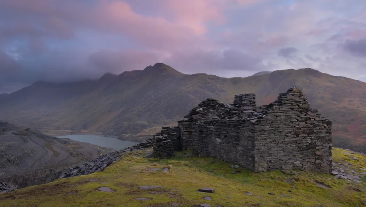 Ruined stone cottage on grassy hillside with mountains and water under dramatic twilight sky.