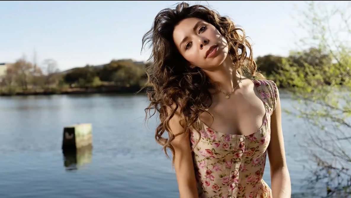 Woman in floral dress gazing upward beside a calm river with tree-lined banks.