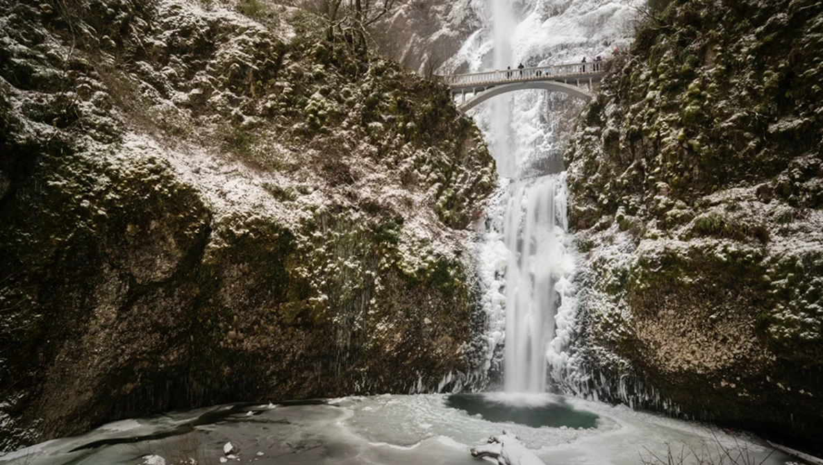 Frozen waterfall cascading between snow-covered canyon walls with a footbridge crossing above.