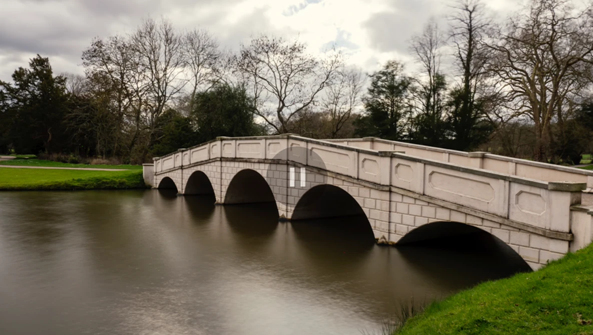 Stone arch bridge spanning a calm river with bare trees and green lawn in overcast daylight.