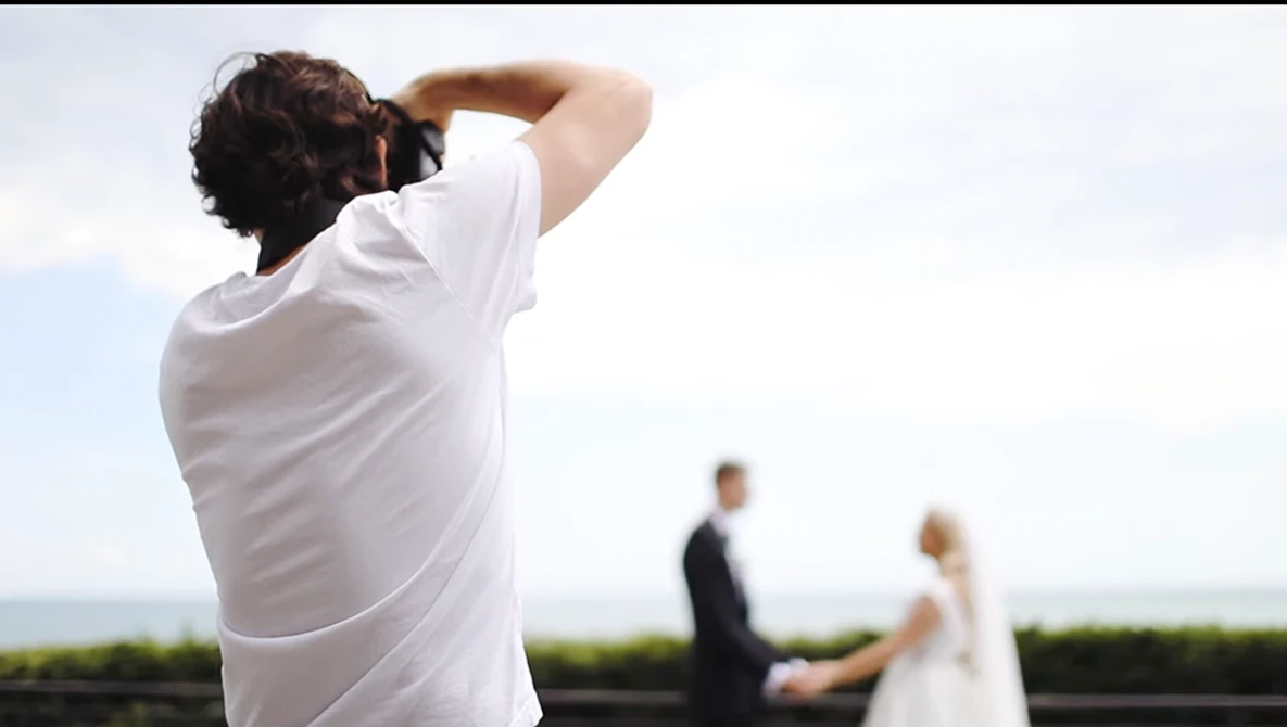 Photographer in white shirt capturing a wedding couple with camera held to face, bride and groom visible in soft-focus background.
