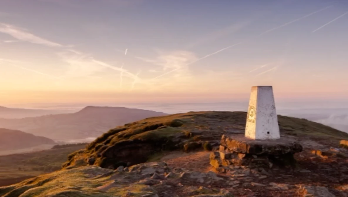 White trig point beacon on a grassy hilltop at golden hour sunrise, with layered valleys and mountains in misty background.