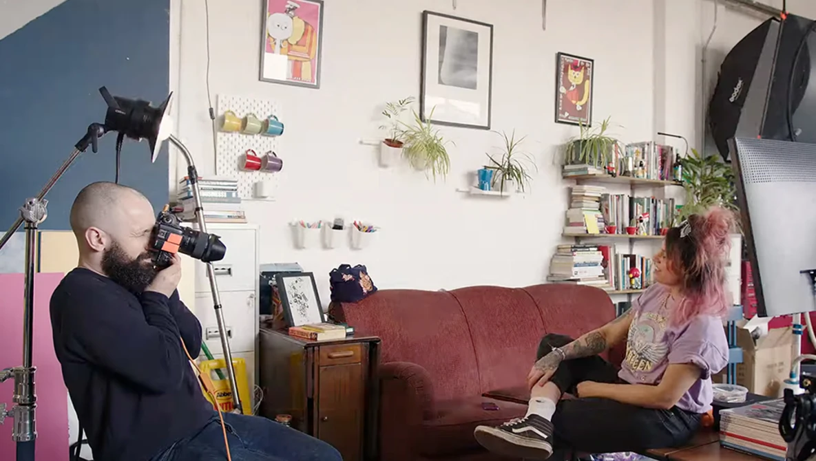 Filmmaker operating camera on tripod while interviewing subject seated on burgundy couch in home studio.